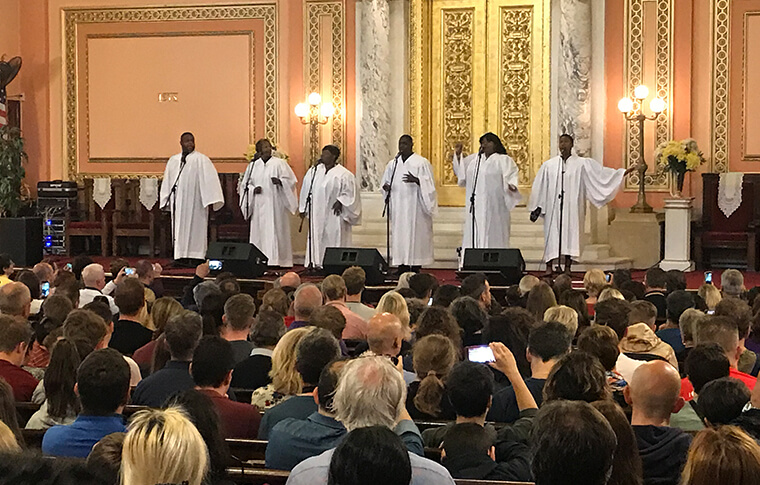 Gospel group on the stage in bright white robes singing into the microphones in front of a large church crowd